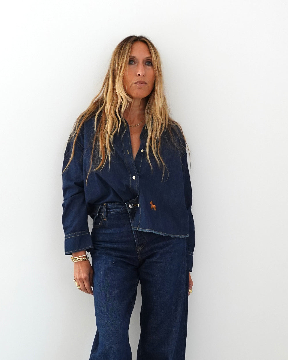 A woman with long blonde hair stands against a plain white wall wearing a dark blue denim button-up shirt and matching dark blue denim jeans.