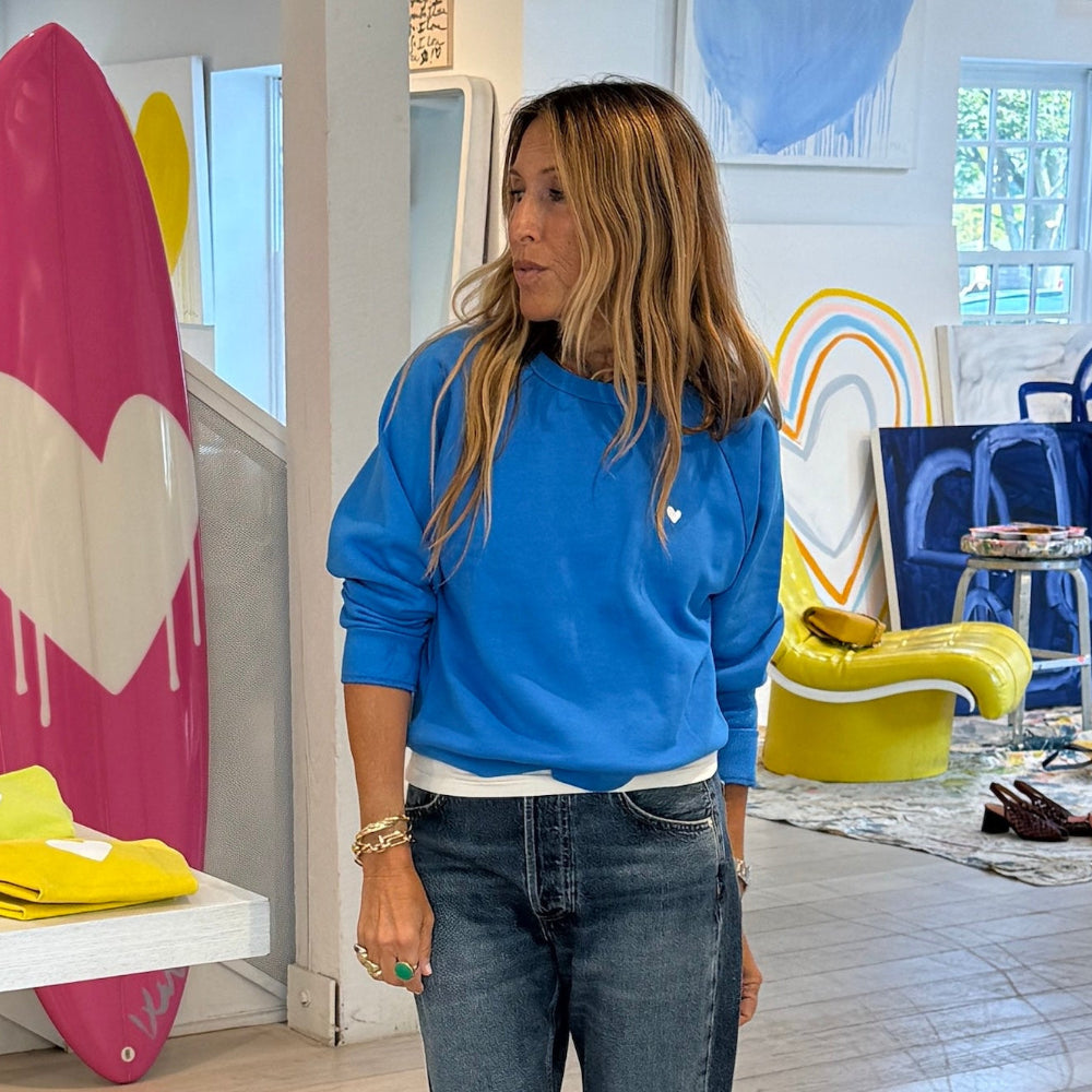 A woman with long light brown hair stands in a brightly lit art studio wearing a bright blue crewneck sweatshirt and dark wash jeans. A large pink surfboard with a white heart is visible on the left, and colorful abstract paintings decorate the background.