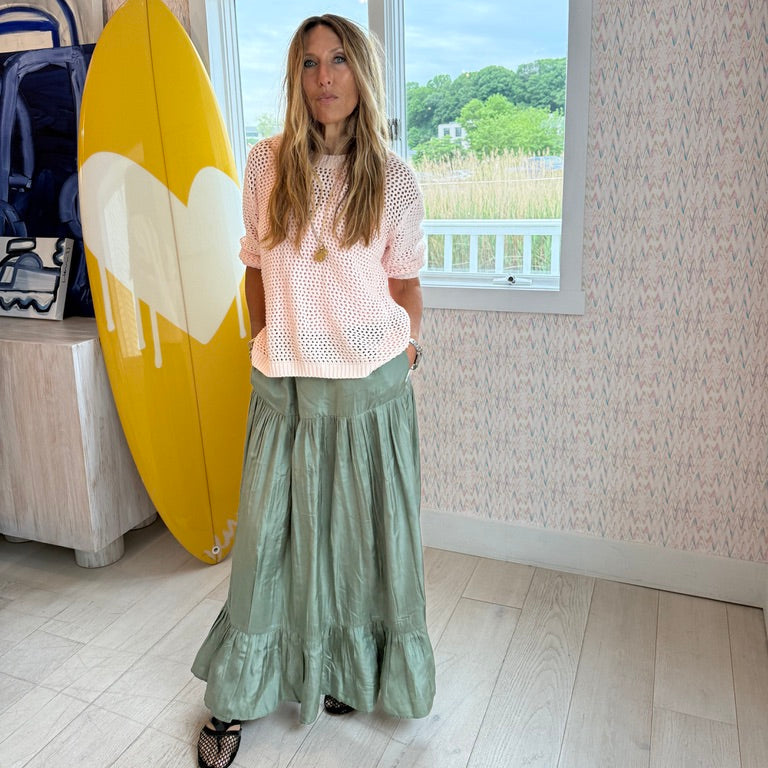 Kerri Rosenthal wearing a pink open-knit crochet sweater and sage green tiered maxi skirt with black heeled sandals, standing in front of a yellow surfboard with a white heart graphic.